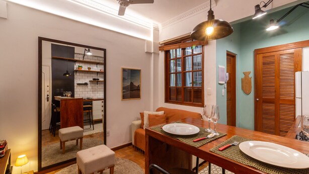 A dining area with a wooden table set for two, a sofa, and a kitchen in the background.