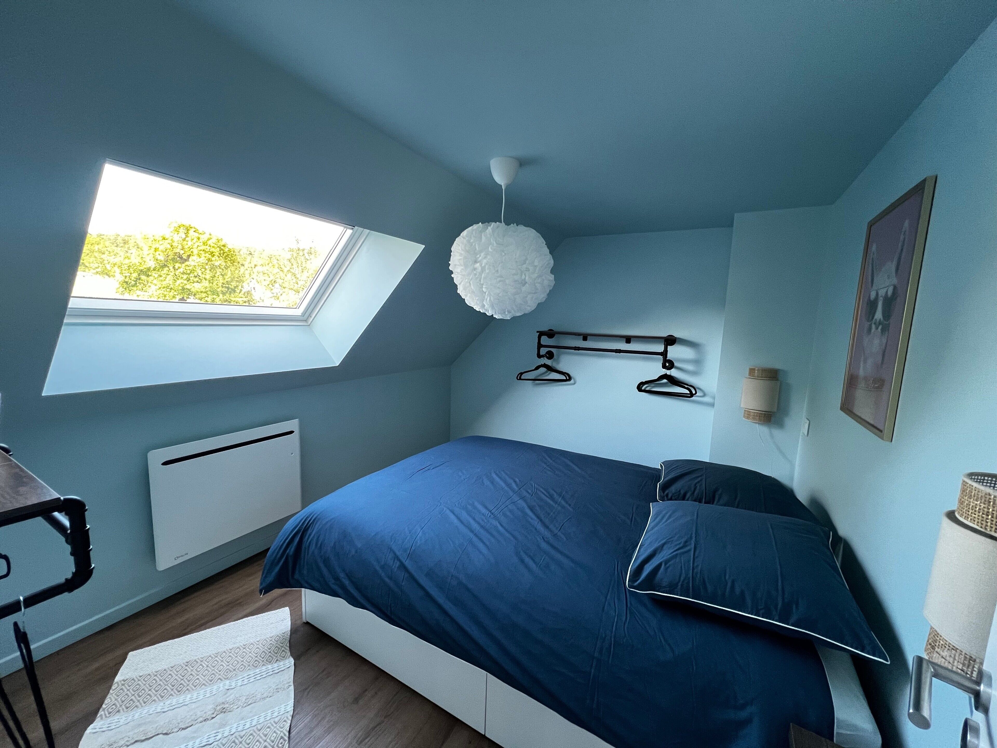 Une chambre avec un puits de lumière, un lit avec une literie bleue, un radiateur blanc, un luminaire suspendu et un tableau encadré au mur.