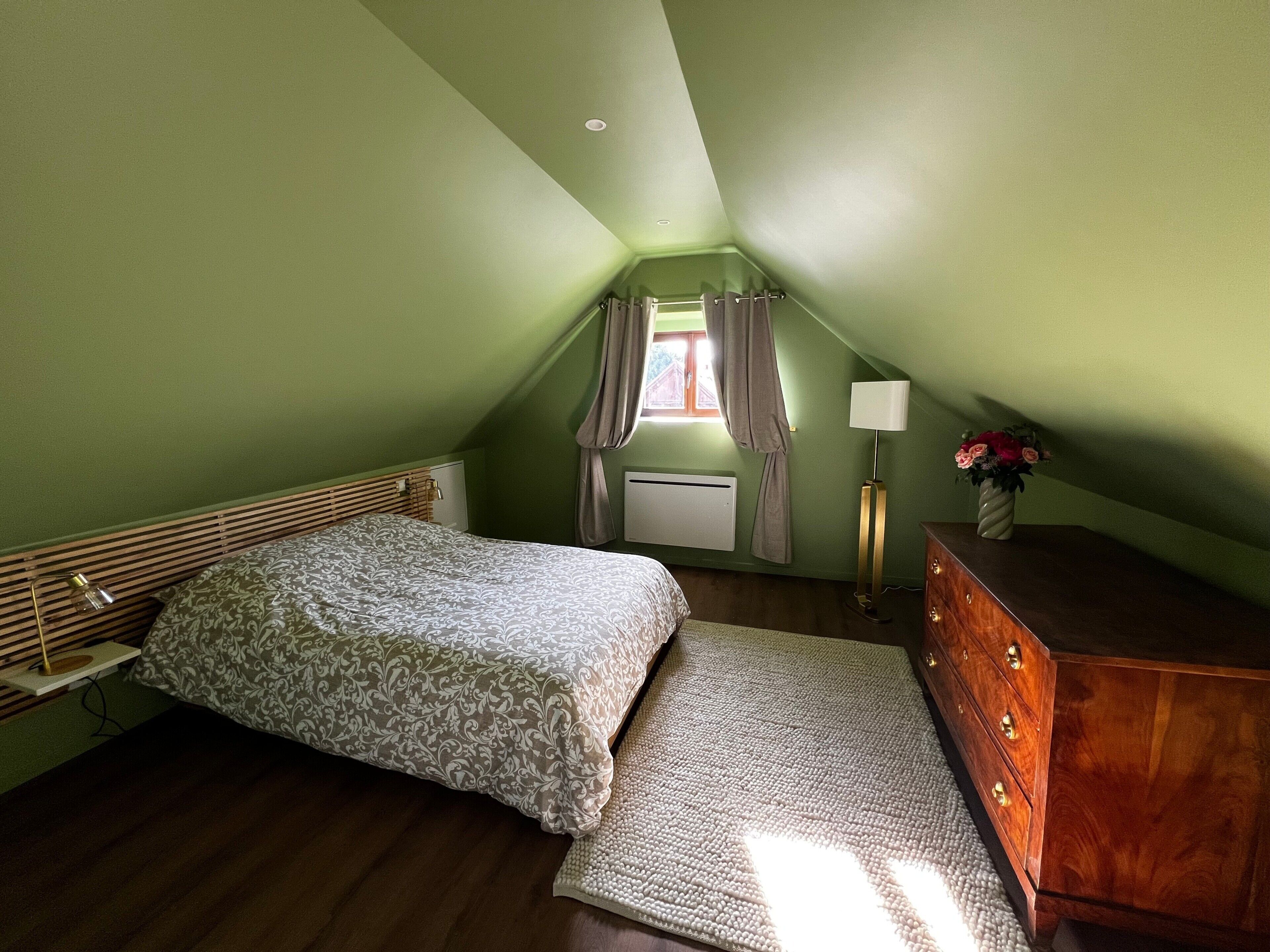 Une chambre à coucher avec un lit, une commode, une lampe et une fenêtre avec des rideaux.