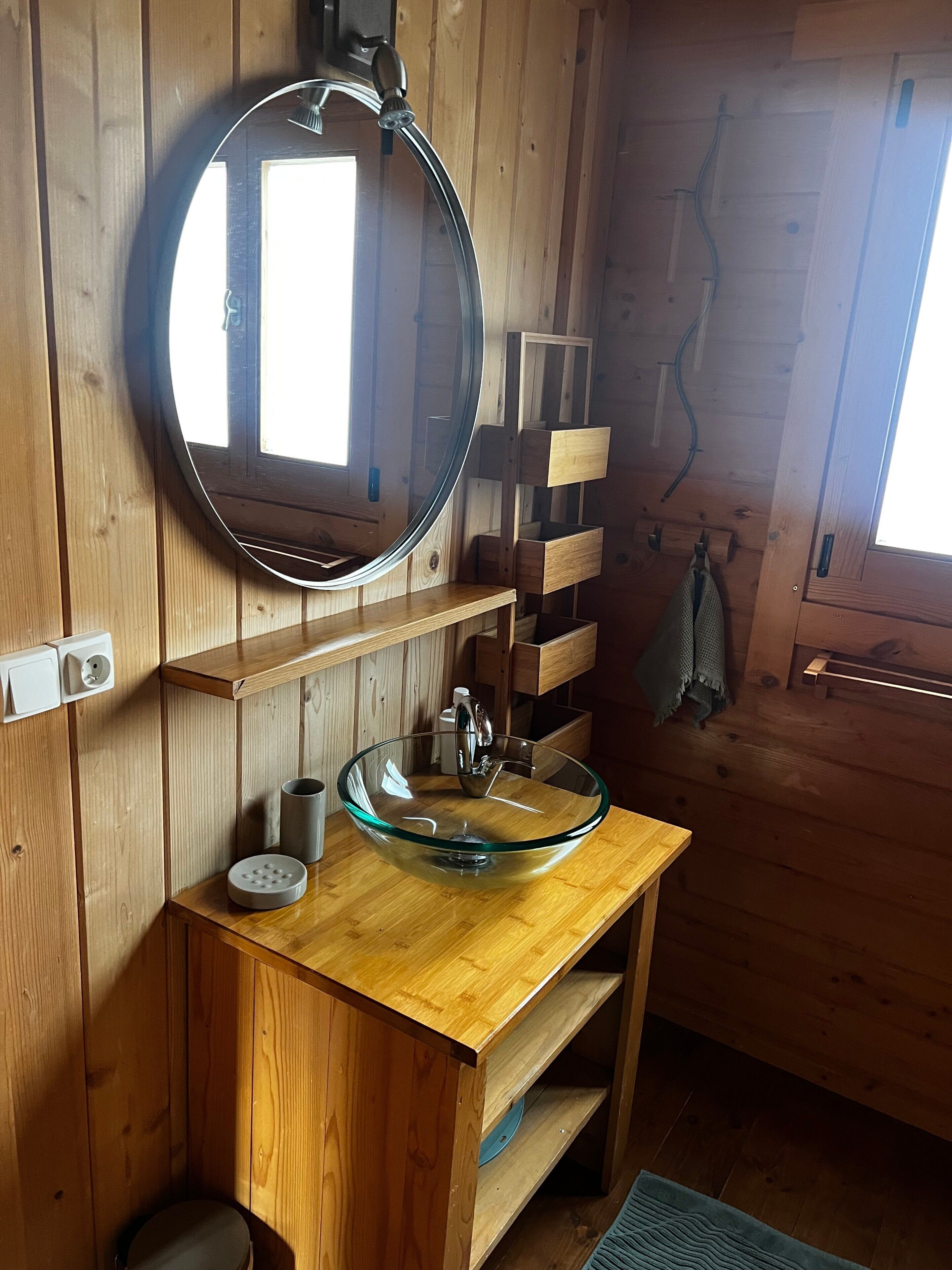 Une salle de bain rustique, avec des murs en bois, un miroir rond, un lavabo en verre, des étagères en bois et une serviette suspendue à un crochet.