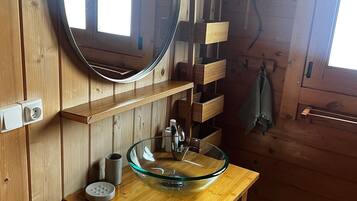 Une salle de bain rustique, avec des murs en bois, un miroir rond, un lavabo en verre, des étagères en bois et une serviette suspendue à un crochet.