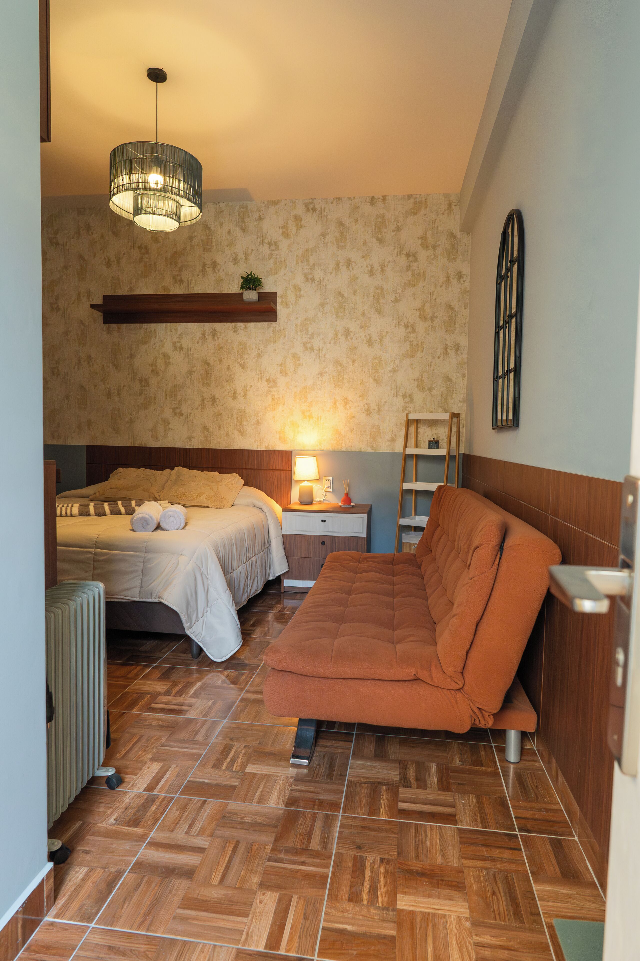 Un dormitorio con cama, sofá, mesita de noche, lámpara, radiador e iluminación colgante.