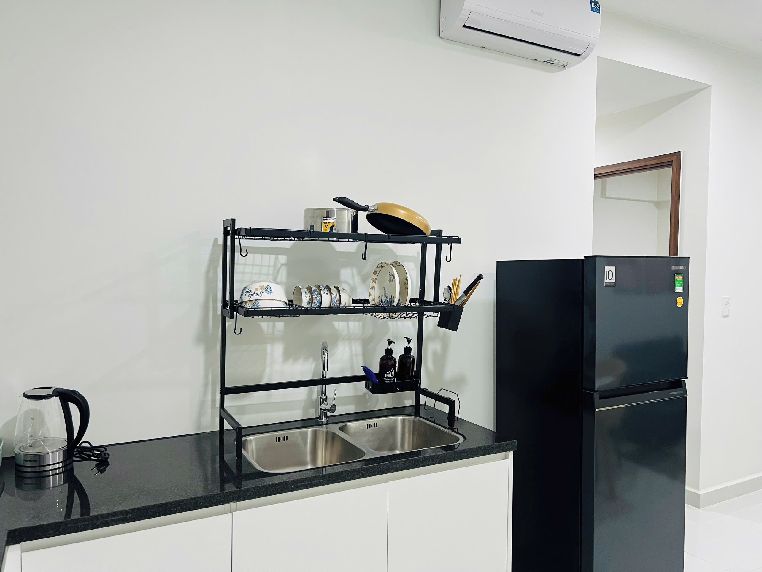A kitchen with a black countertop, a double sink, a black refrigerator, and a dish rack with dishes and utensils.