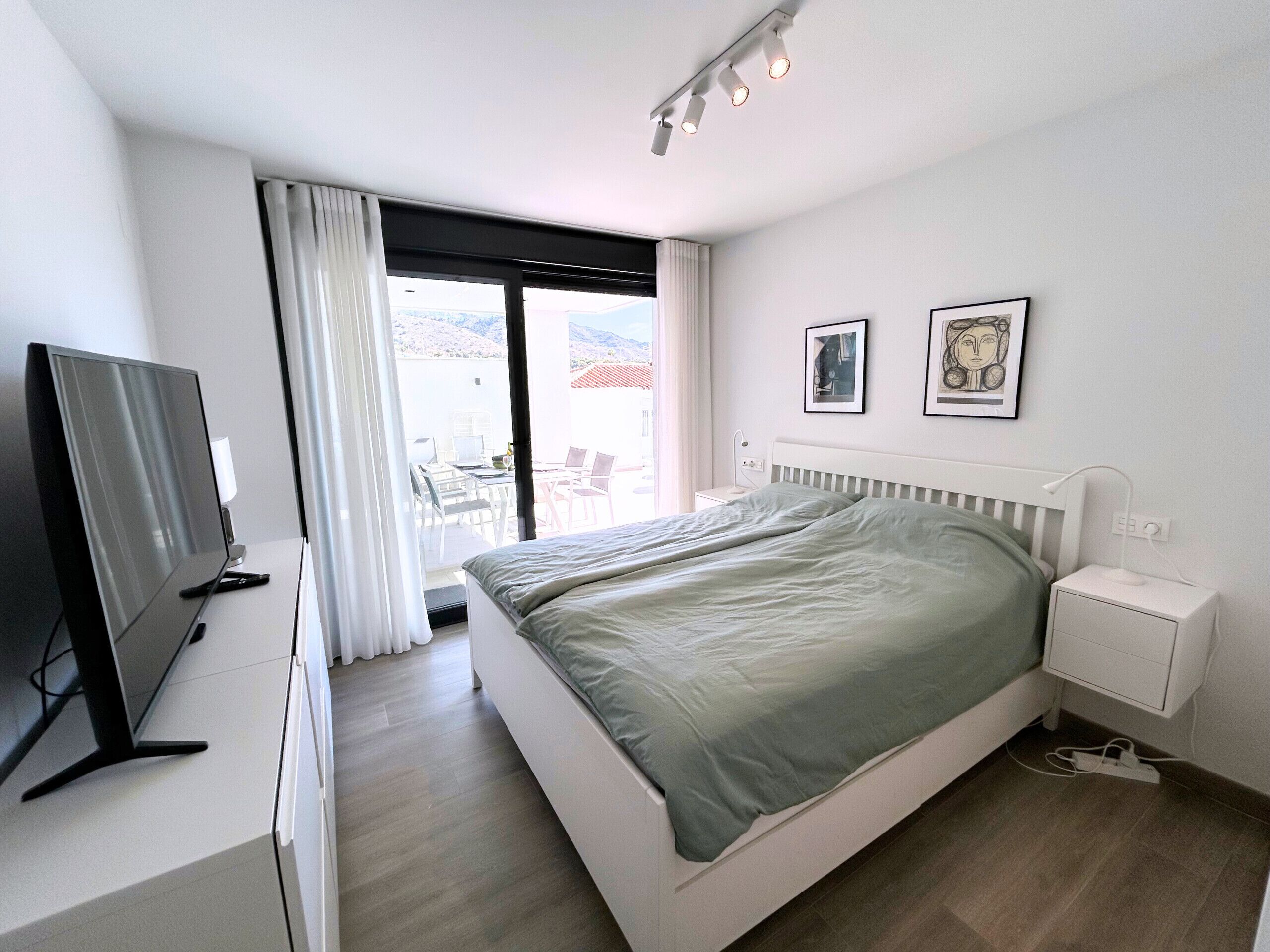 A modern bedroom with a bed, bedside table, TV, and a balcony with outdoor seating.