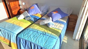 A room with two beds, each with a blue and yellow quilt, pillows, and rolled towels. There is a wooden nightstand with a lamp between the beds. - Room + bathroom, splendid sea view, well equipped (Callao Salvaje)