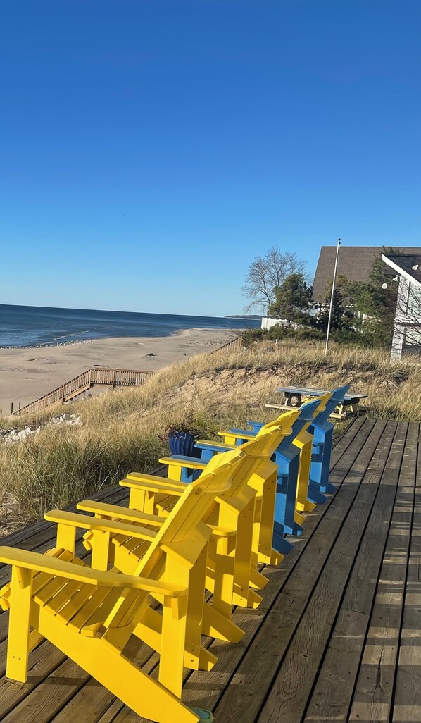 Ett trädäck med gula och blå Adirondack-stolar, placerat med utsikt över en strand.