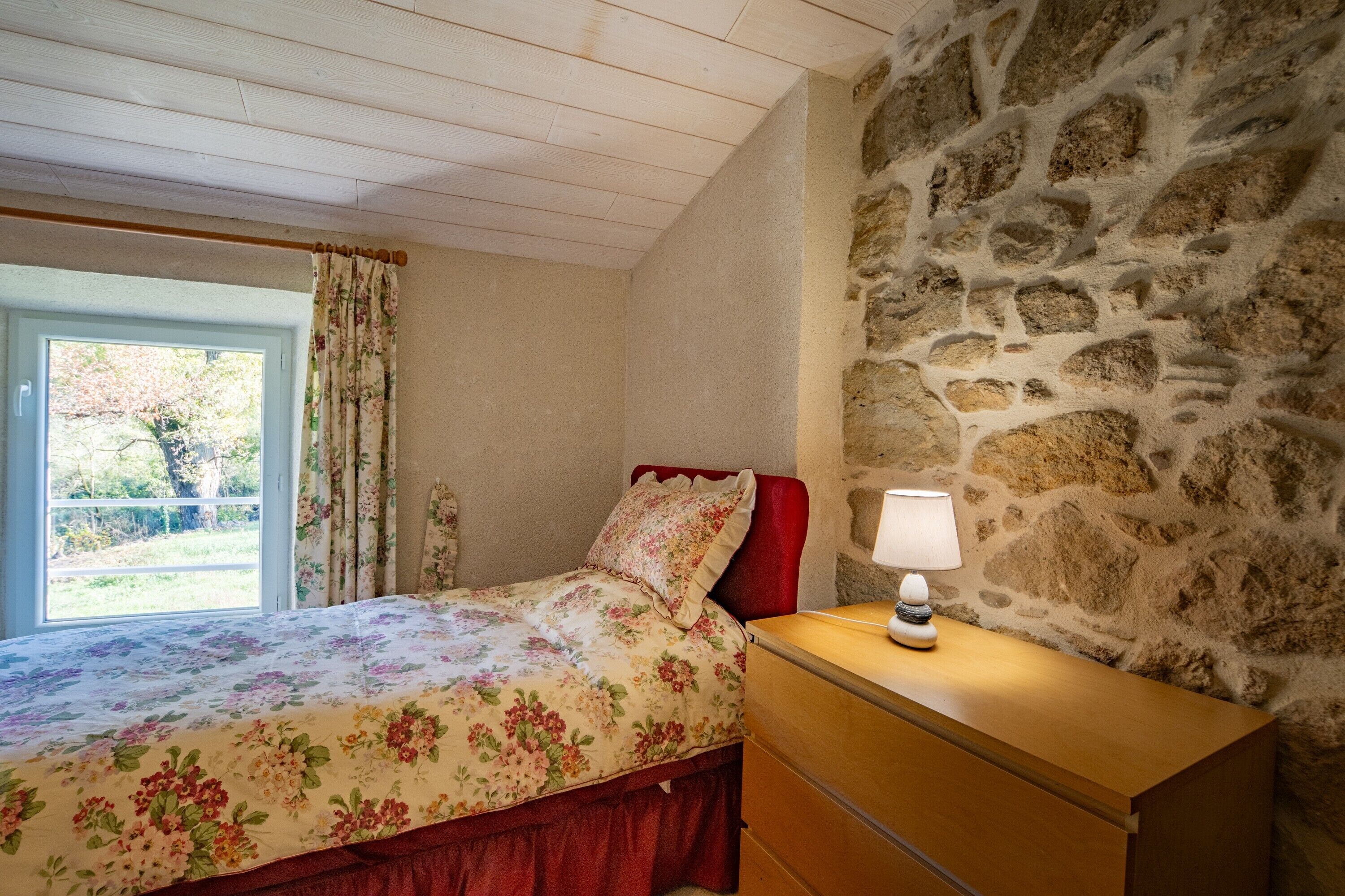 A bedroom with a stone wall, a wooden bed with floral bedding, a bedside table with a lamp, and a window with curtains.