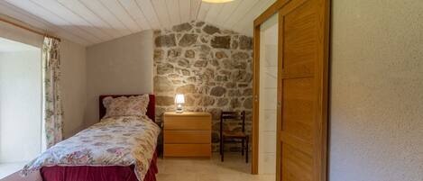 Un dormitorio con pared de piedra, armario de madera, mesita de noche con lámpara, una silla y ventana con cortinas.