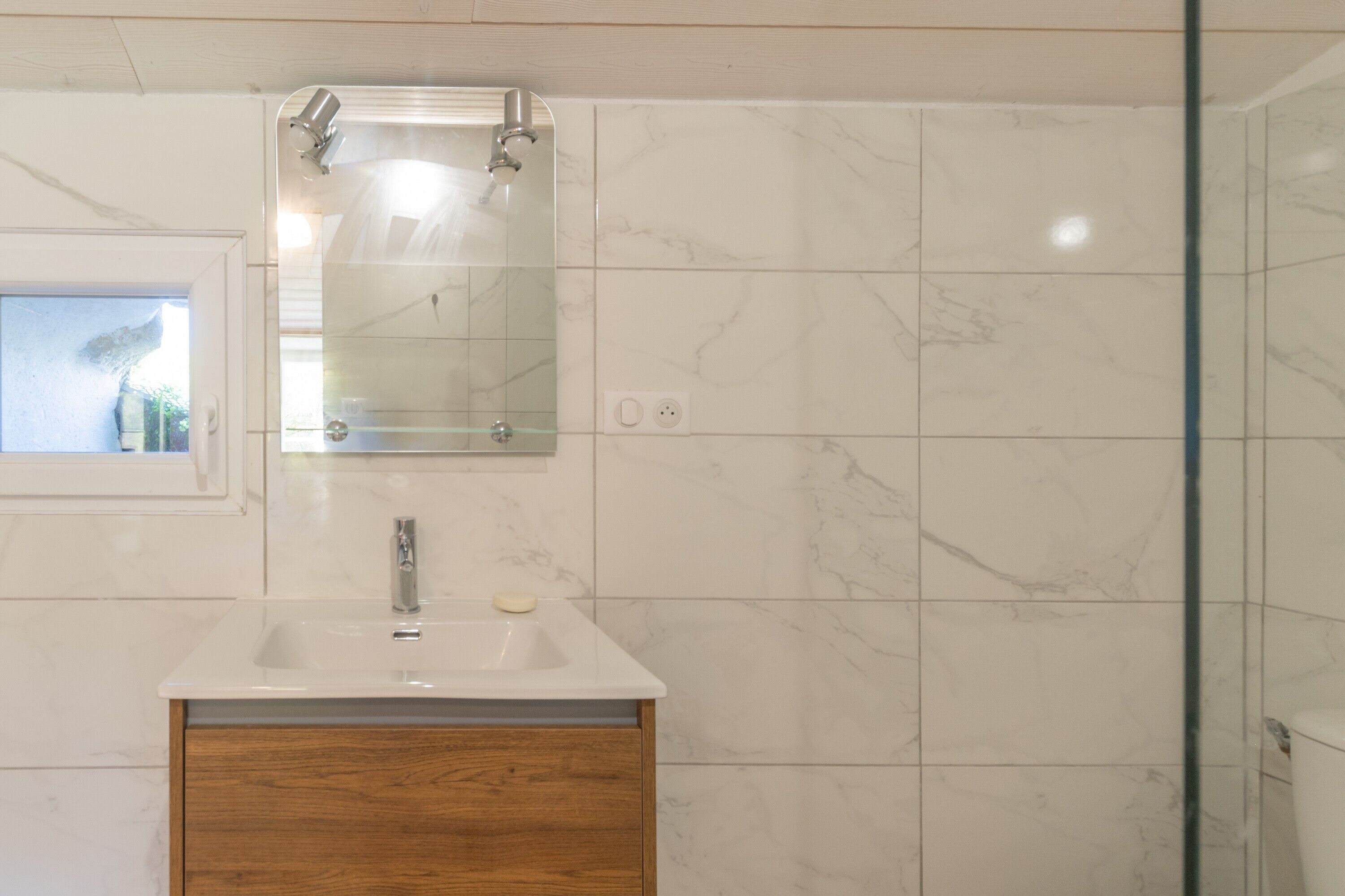 A bathroom with a white sink, wooden cabinet, marble tiles, a mirror, and a shower.