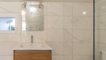 Um banheiro com pia branca, armário de madeira, azulejos de mármore, um espelho e um chuveiro.