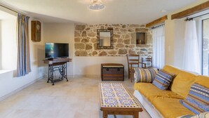 A living room with a stone wall, a television, a wooden table, a sofa, and a patterned tablecloth. - Cottage 'L'Ancienne Estable' with Private Terrace, Private Garden and Air Conditioning (Les Assions)