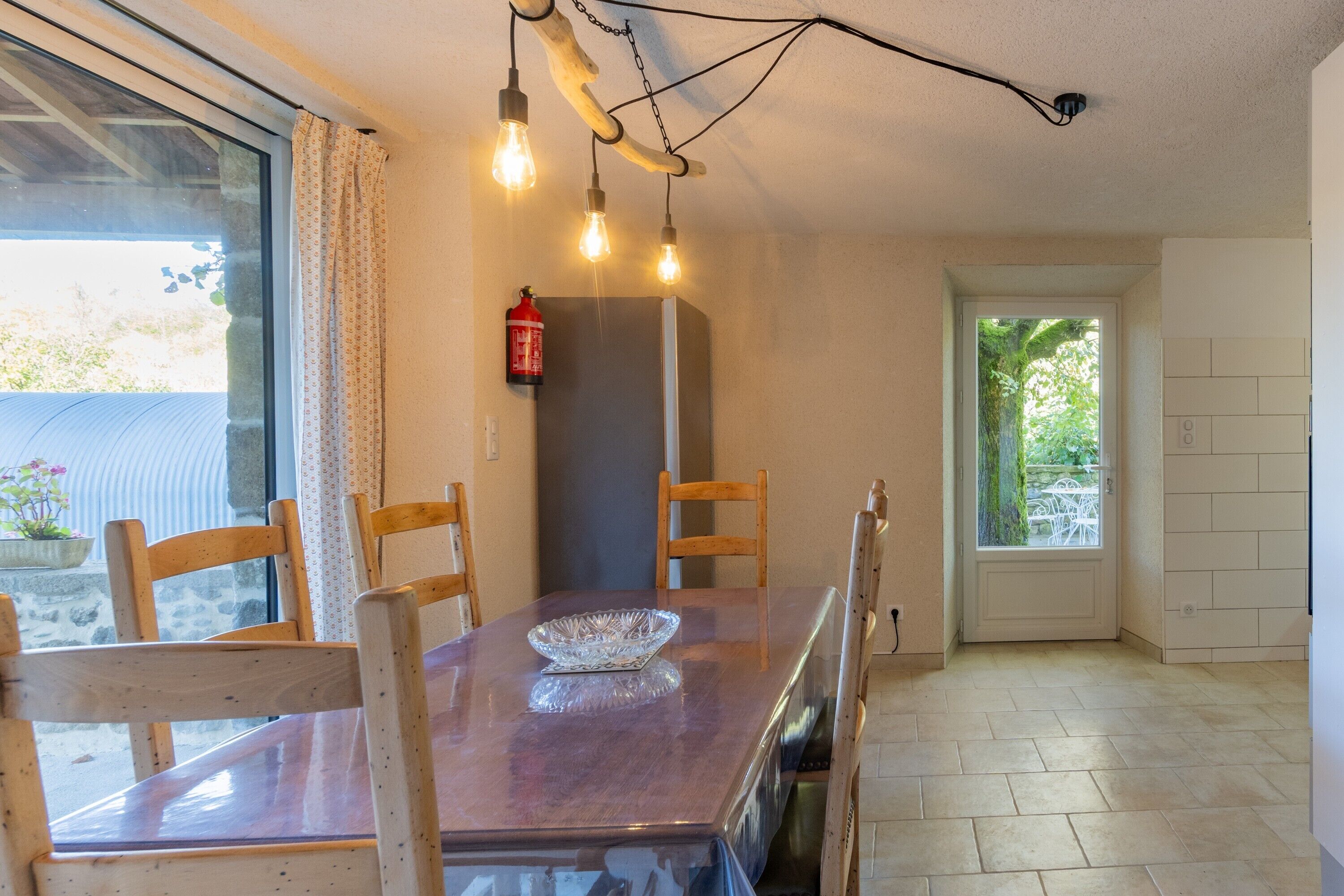 A dining area with a wooden table, chairs, and a glass bowl. There are hanging light bulbs and a fire extinguisher on the wall.