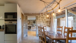 A kitchen with modern appliances, a dining area with wooden chairs, and a stone wall. - Cottage 'L'Ancienne Estable' with Private Terrace, Private Garden and Air Conditioning (Les Assions)