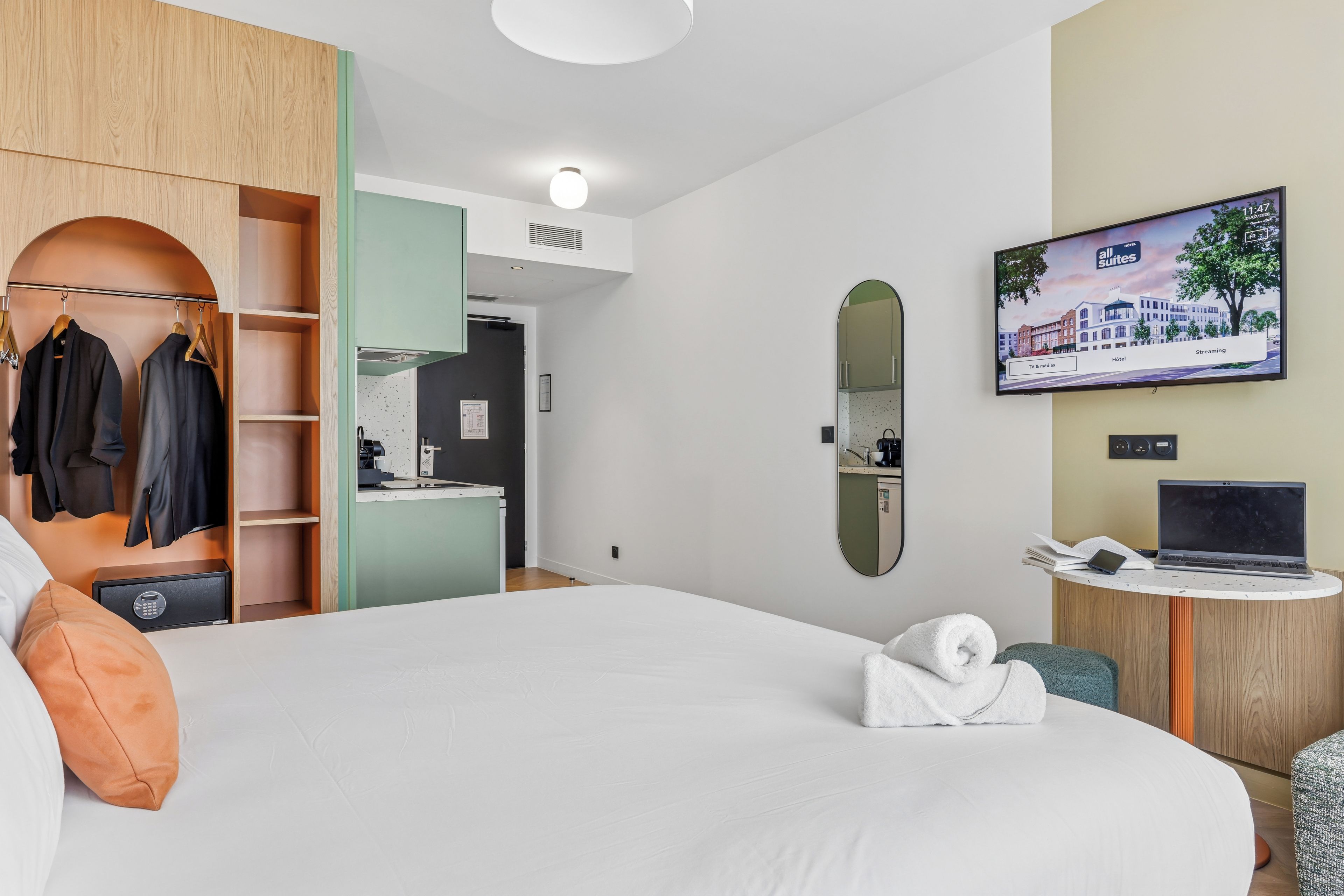 A hotel room with a bed, a wardrobe, a desk with a laptop, a TV, and a mirror.