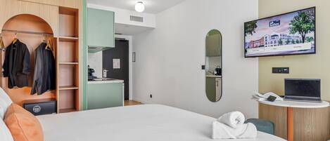 A hotel room with a bed, a wardrobe, a desk with a laptop, a TV, and a mirror.