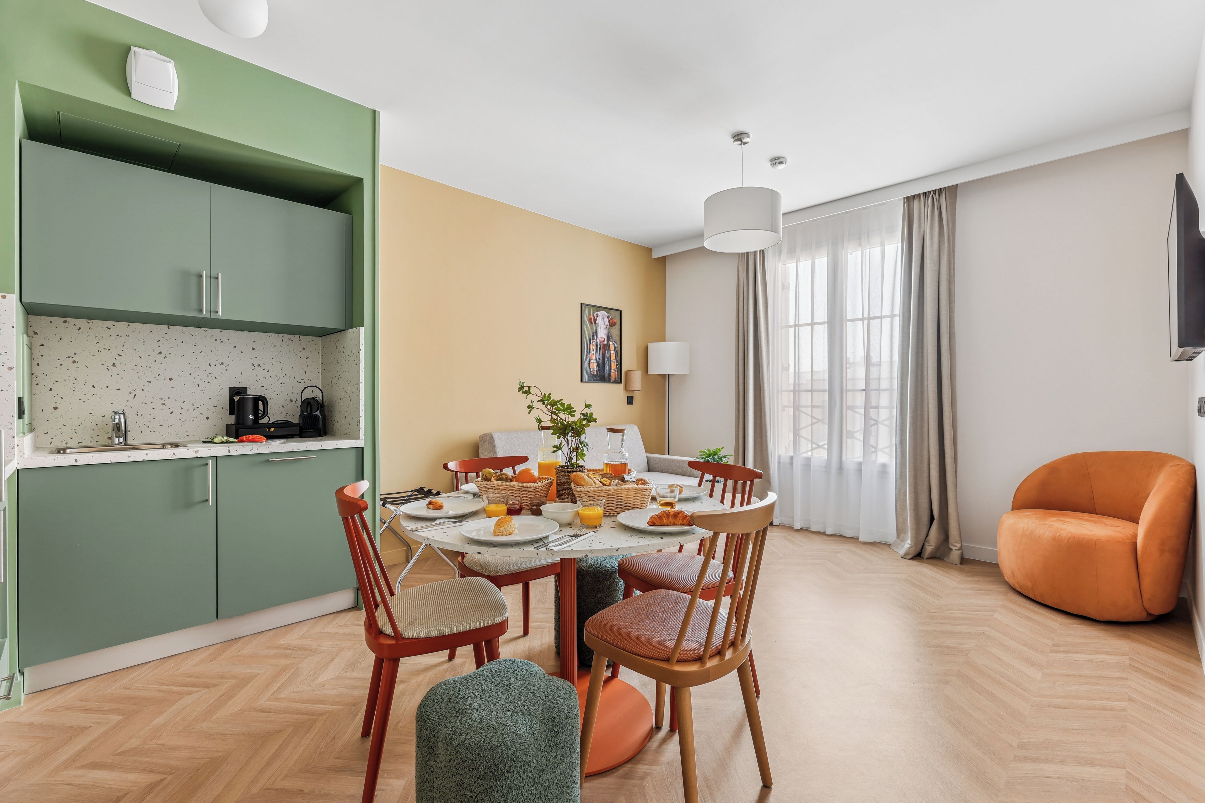 A modern kitchen with green cabinets, a dining area with a round table, and a comfortable orange chair.