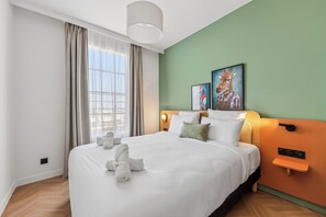 Quarto de hotel com uma cama grande, cabeceira com luminária, duas fotos emolduradas na parede e uma janela com cortinas.
