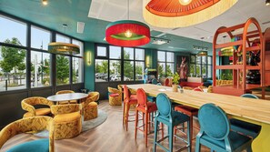 A modern interior with a wooden table, colorful chairs, and large windows offering a view of the street. - All Suites Appart Hôtel | Val d’Europe près de Disneyland® Paris (Serris)