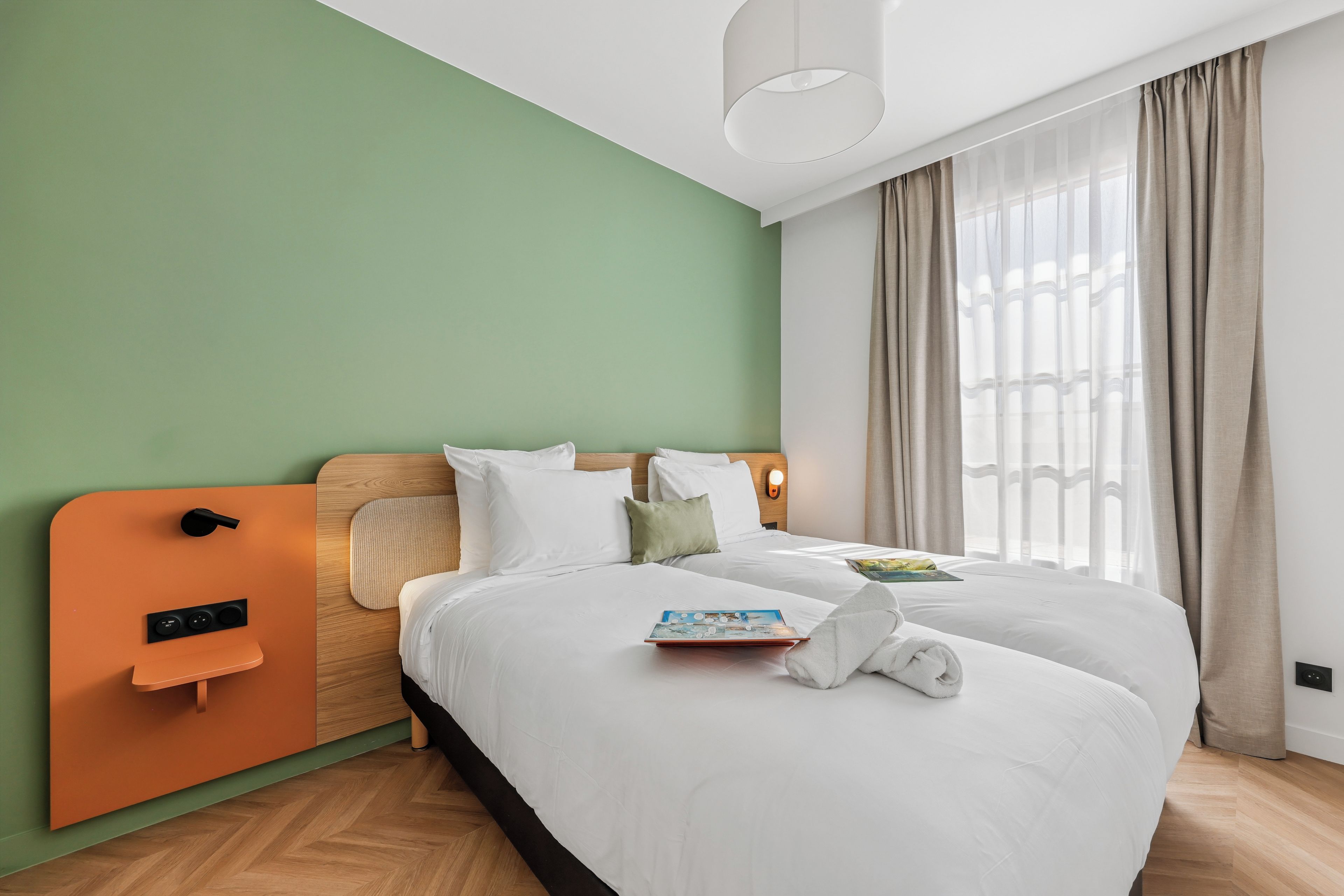 Habitación de hotel con cama, mesita de noche, panel de control en la pared y ventana con cortinas.