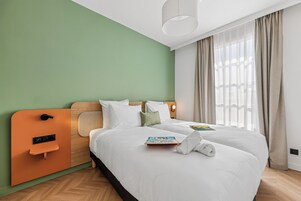 Habitación de hotel con cama, mesita de noche, panel de control en la pared y ventana con cortinas.