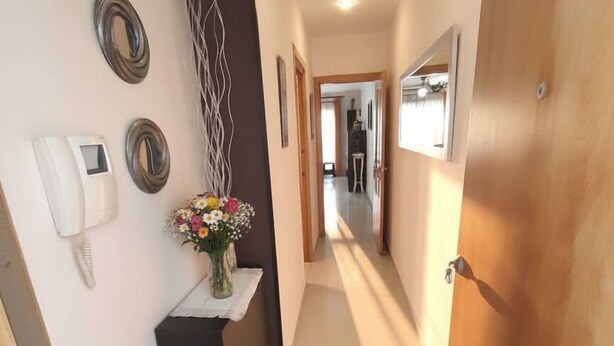 Un pasillo estrecho con una puerta de madera a la derecha, una pared blanca con un teléfono y espejos decorativos, y una pequeña mesa con un jarrón de flores.