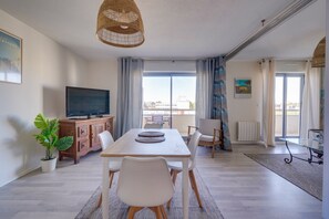 A spacious living area with a wooden dining table, white chairs, a flat-screen TV, and a large window offering a city view. - Le Charme D'Antan - quartier Chartrons (Bordeaux)