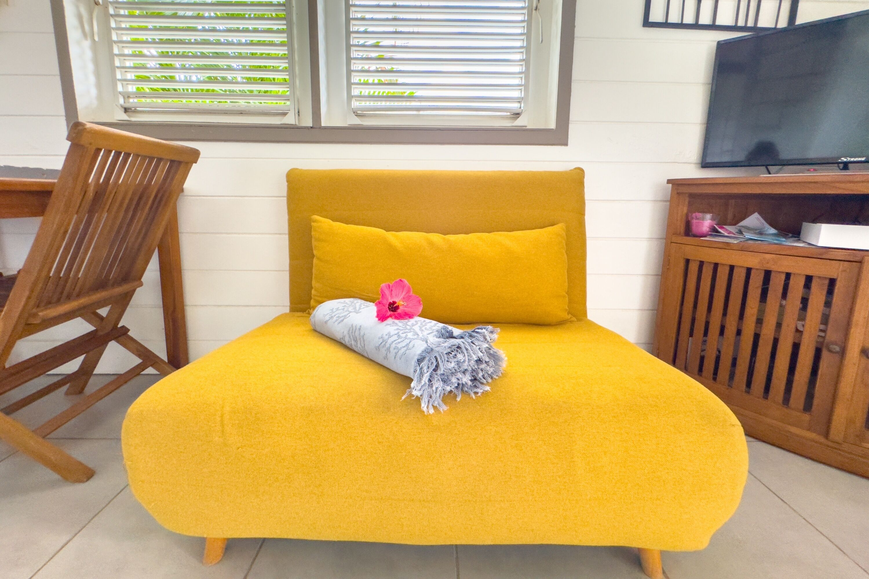 Un sofá amarillo con un cojín y una manta, una silla de madera, un televisor y una ventana con persianas.