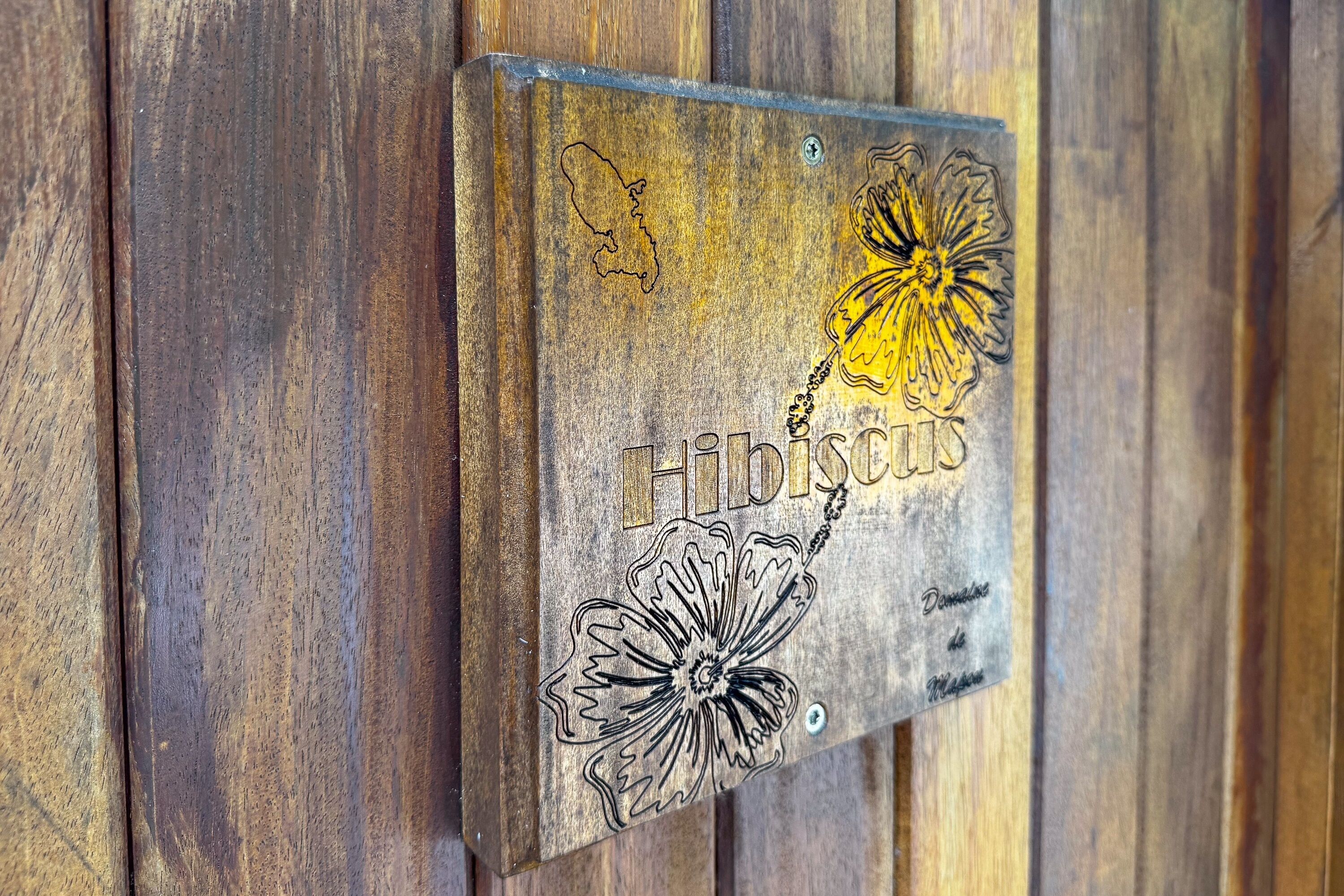 A metal plaque with a flower design and the name "Hibiscus" engraved on it, mounted on a wooden wall.