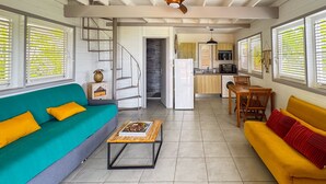 A spacious living area with a sofa, a coffee table, and a dining table. A kitchen is visible in the background with a microwave and a refrigerator. - Lodge 2 - Hibiscus - Jaccuzi Privé (Les Anses-d'Arlet)