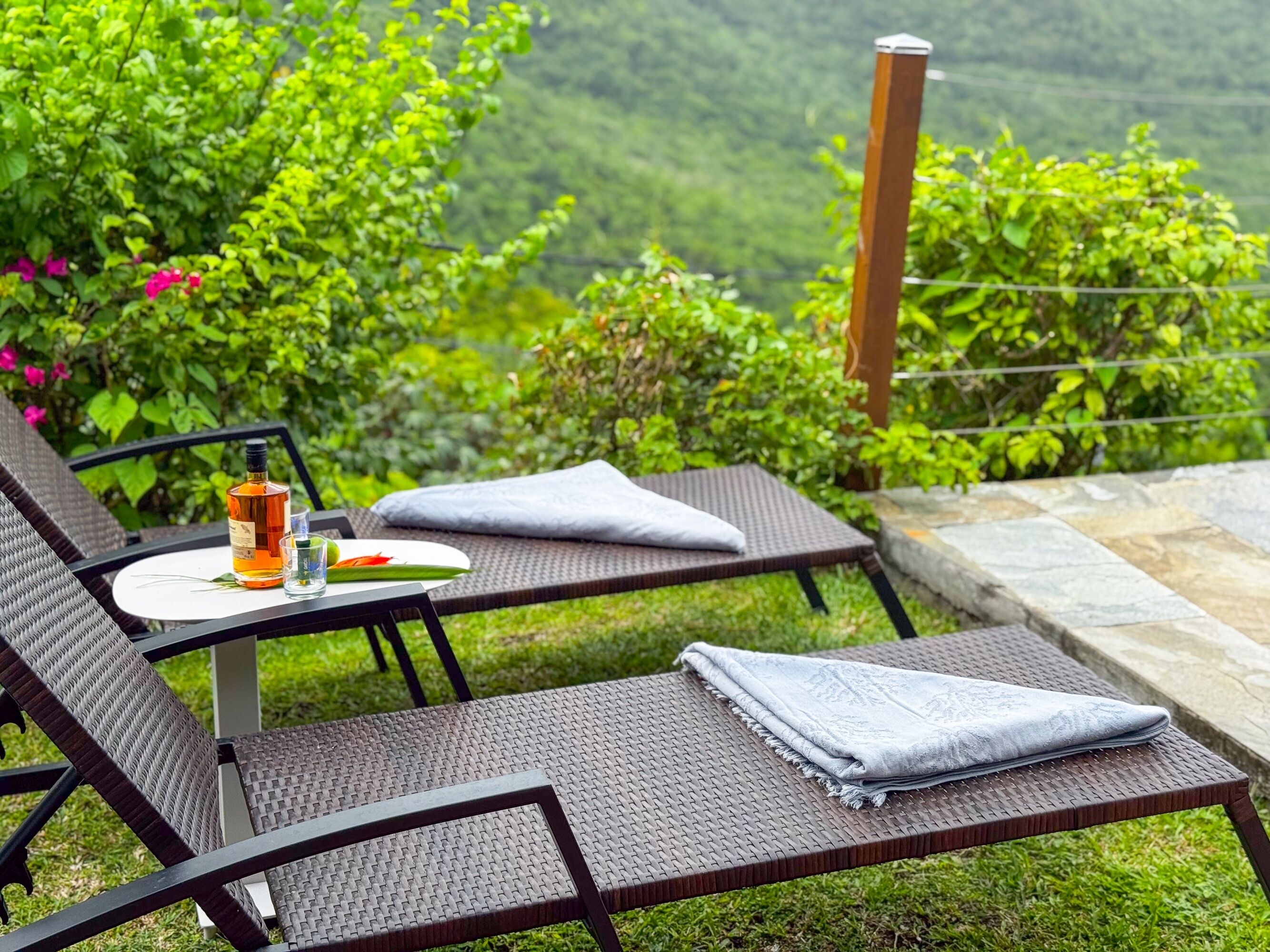 A patio with two wicker lounge chairs, a table with a bottle and glasses, and a view of lush greenery.