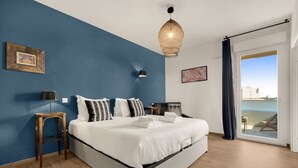 A modern bedroom with a large bed, a wooden nightstand, a floor lamp, and a view of the sea through a sliding glass door.