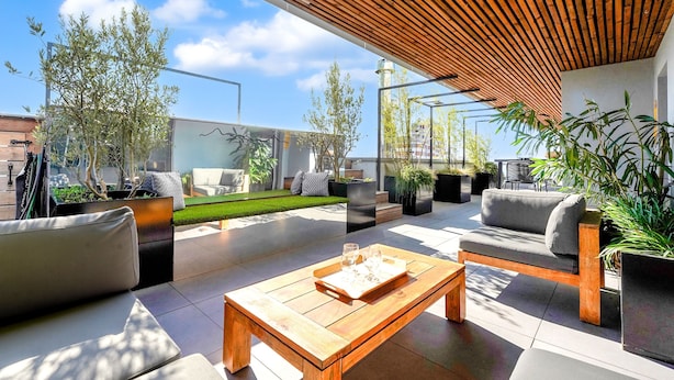 An outdoor patio area with modern furniture, including a wooden table with a tray, a grey sofa, and a wooden bench. There are potted plants and a clear view of the sky.