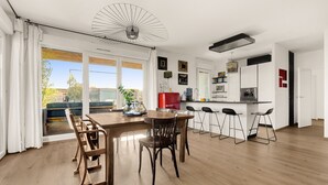 A modern kitchen with a dining area, featuring a wooden table and chairs, a bar area with stools, and a large window with curtains.