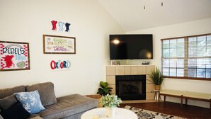 Een woonkamer met een grijze bank, een ronde salontafel en een flatscreen televisie boven een open haard.