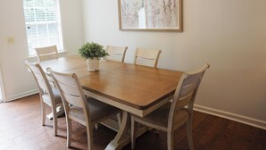 Een eetkamer met een houten tafel en zes stoelen, een plant in pot, een kroonluchter en een ingelijst botanisch kunstwerk aan de muur.
