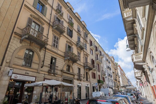 Une rangée de bâtiments historiques dotés de balcons, un restaurant appelé « La Piola Sarauda » situé au rez-de-chaussée, et des voitures garées le long de la rue.