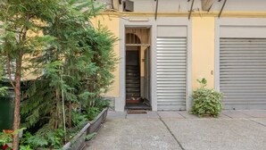 Un étroit chemin de béton mène à l’entrée d’un bâtiment, qui est dotée d’une porte en métal et d’un petit seuil.
