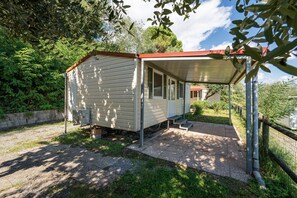 A small, single-story building with a covered patio area, surrounded by greenery and a gravel path. - Mobile Home 'Camping Belsito Dalmacija 505' with Shared Pool, Wi-Fi and Air Conditioning (Vico)