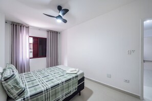 A bedroom with a bed, curtains, a ceiling fan, and a window. - Apartment 90 meters from the beach + free garage and amenities (Praia Grande)