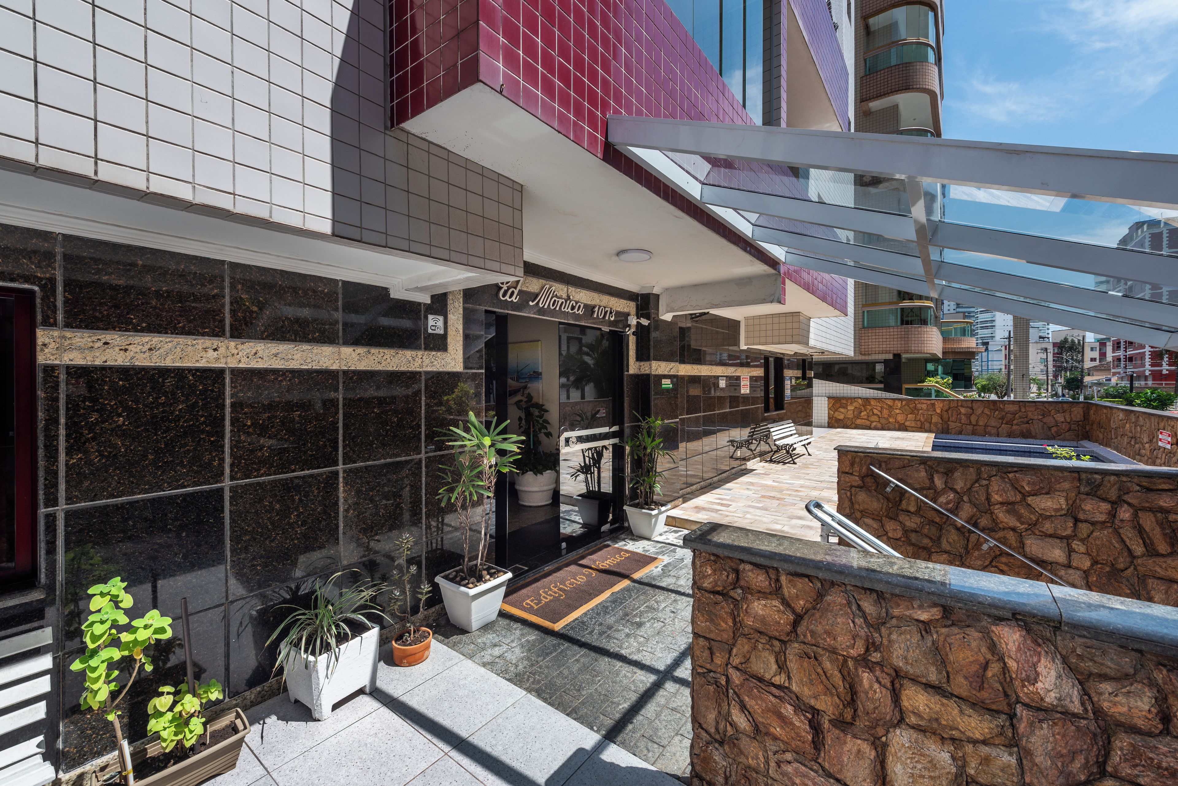 A hotel entrance with a modern design, featuring a tiled facade, a stone wall, and a small outdoor seating area with potted plants.