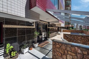 A hotel entrance with a modern design, featuring a tiled facade, a stone wall, and a small outdoor seating area with potted plants.