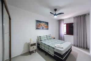 A bedroom with a bed, a nightstand, a lamp, a ceiling fan, a window with curtains, and a wall-mounted painting of a tree.