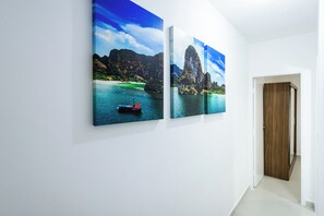 A hallway with two wall-mounted paintings of a tropical landscape featuring a boat and a large rock formation. - Apartment 90 meters from the beach + free garage and amenities (Praia Grande)