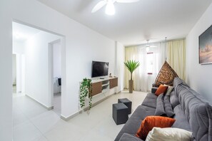 A modern living room with a grey sofa, a flat-screen TV, a potted plant, and a hanging chair. - Apartment 90 meters from the beach + free garage and amenities (Praia Grande)