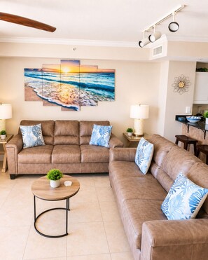 A living room with a brown leather sofa, a coffee table with a plant, and a wall-mounted painting of a beach scene. - Beachfront Convenient Unit #204! (Panama City Beach)