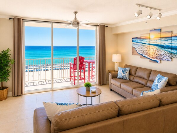 A room with a large sliding glass door leading to a balcony with a view of the ocean. There is a brown leather sofa, a small round table, and a red chair on the balcony.