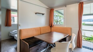 A compact room with a wooden table, orange cushioned bench, white chairs, a small bed, and a window with curtains. - Mobile Home 'Camping Belsito Dalmacija 506' with Shared Pool, Wi-Fi and Air Conditioning (Vico)