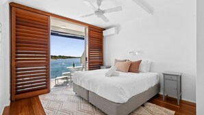 A bedroom with a bed, bedside tables, a nightstand, a ceiling fan, and a view of the sea through large windows. - Mariners Point 5 Noosaville (Noosaville)