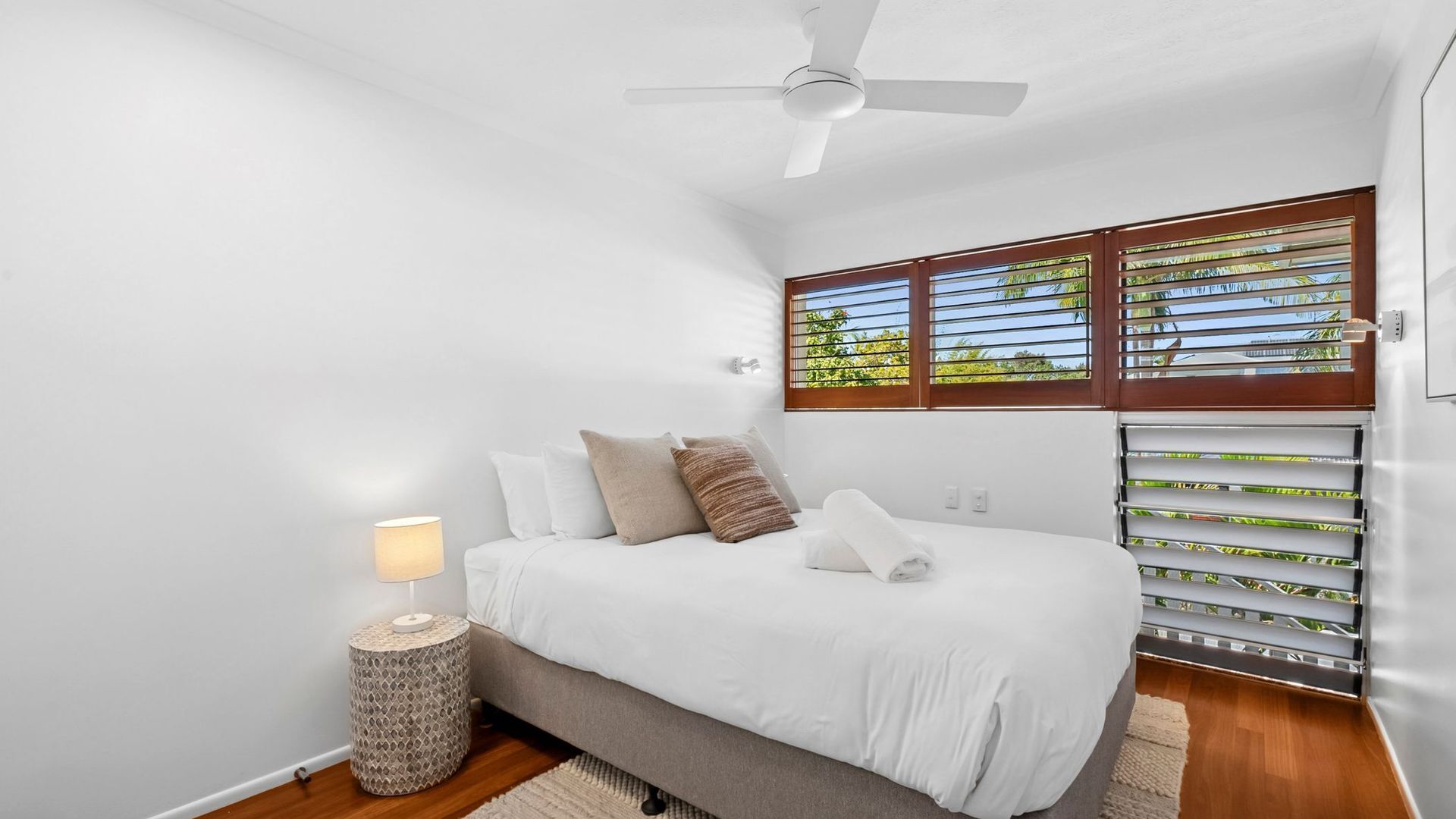 A bedroom with a bed, a ceiling fan, a window with blinds, and a bedside lamp.