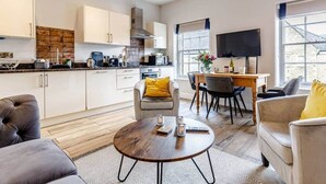 A modern kitchen with white cabinets, a dining area with a wooden table and chairs, and a living room with a sofa and a coffee table.